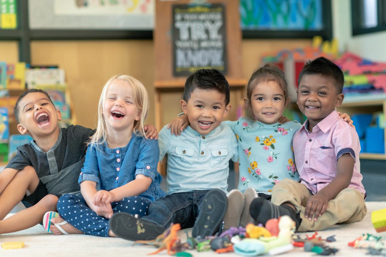 Children sitting in a line with their arms around each other. The kids are laughing and smiling directly at the camera.