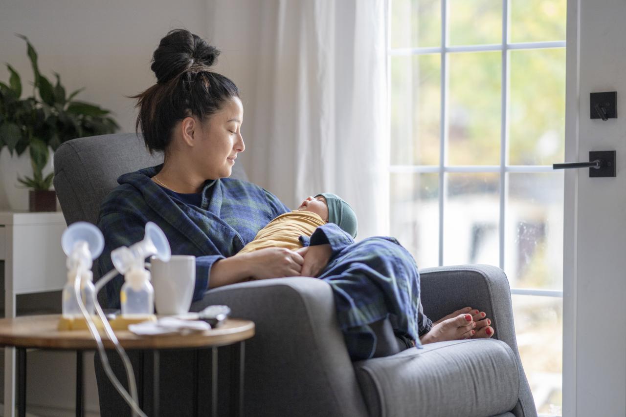A new mother sitting on a rocking chair in her living room wearing a housecoat holding her newborn baby.