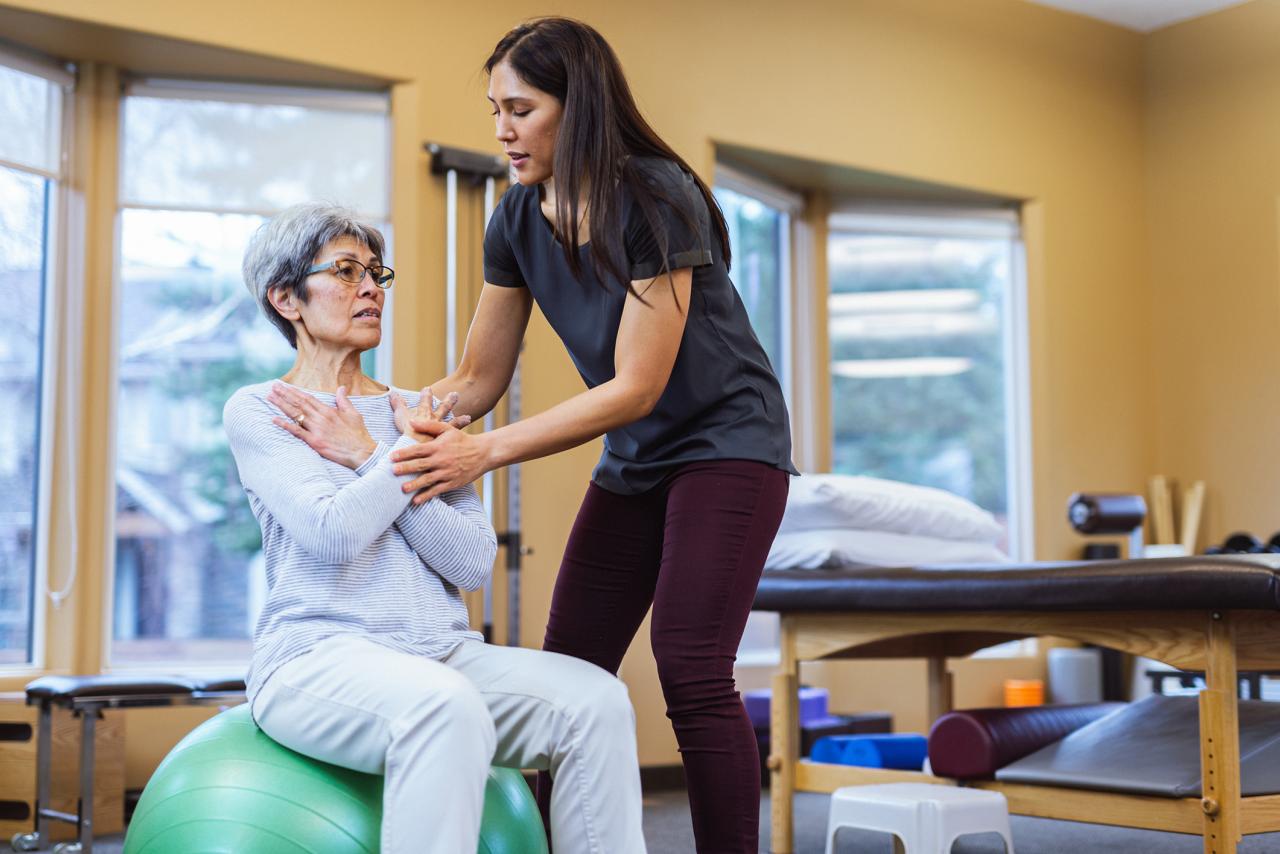 Female sits on a yoga ball, with the help of female physical therapist standing beside her as they do exercises