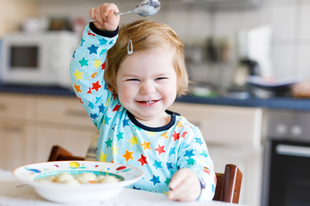 a smiling baby holding up a spoon with bowl of food in front of them.