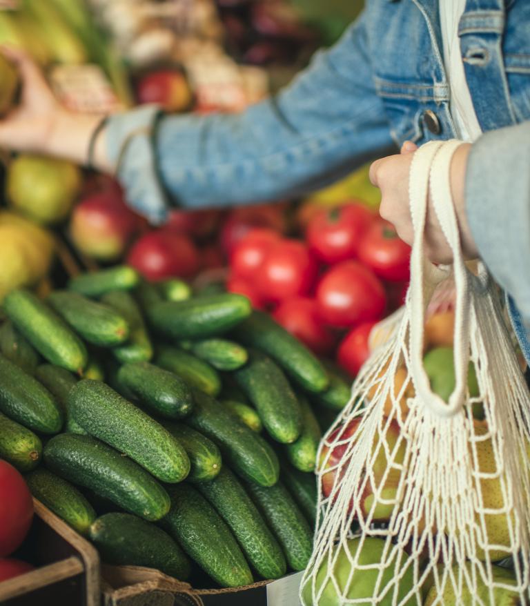 Person at a farmers market selecting fruits and vegetables using a reusable bag