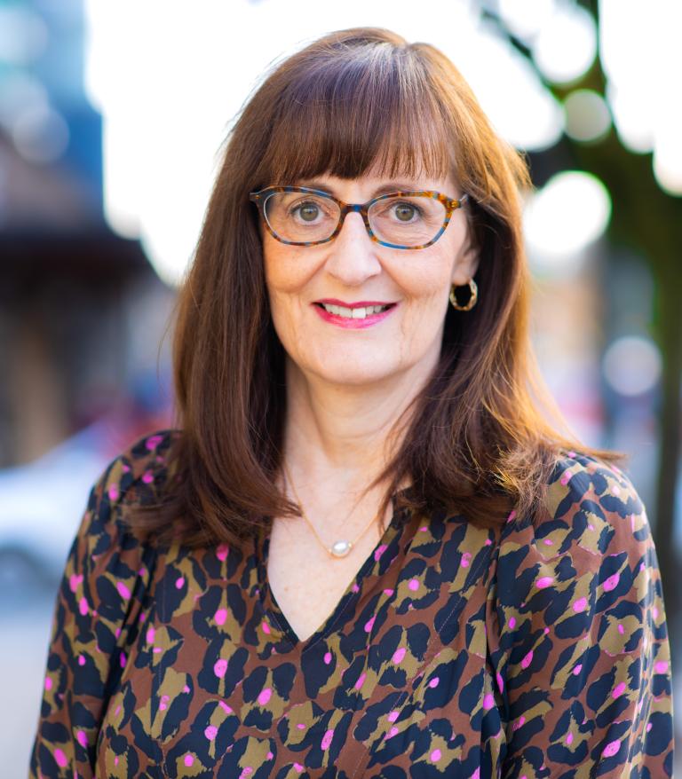 A portrait of Patricia Daly outside on a sunny day. She is glasses and an animal print blouse
