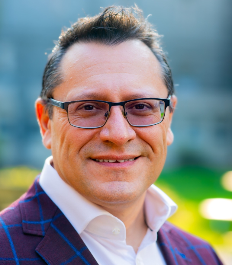 A portrait of Fernando Pica outside on a sunny day. He is wearing a white shirt and purple blazer