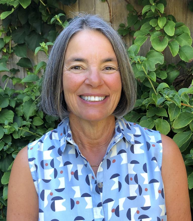 A portrait of Kathy Greenberg in front of green plants. She is wearing a blue and white patterned top