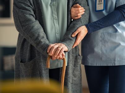 A closeup of a nurse helping an older person with a cane walk down a hallfway