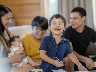 A parent holding a young child while cooking dinner, a young boy is helping stir the pot.